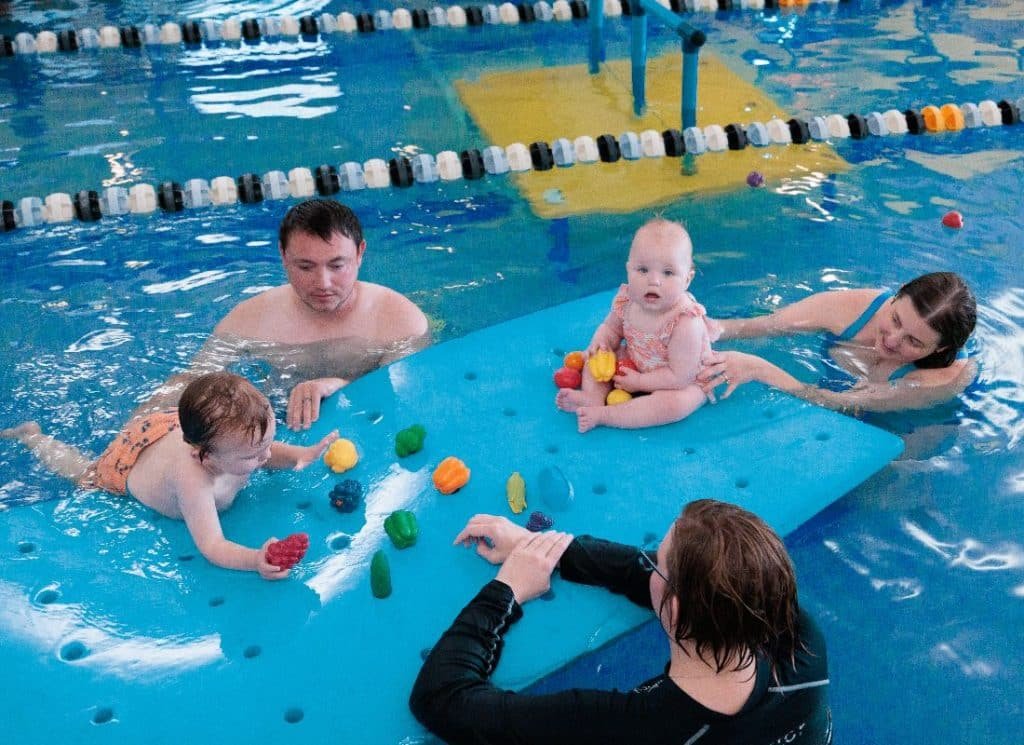 Two adults and two babies play with colorful toys on a blue mat in a swimming pool.