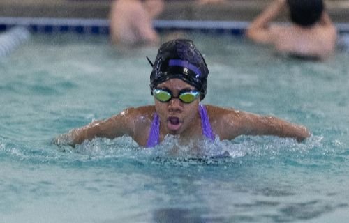 A swimmer in a black cap, green goggles, and purple swimsuit performs the butterfly stroke in a pool.
