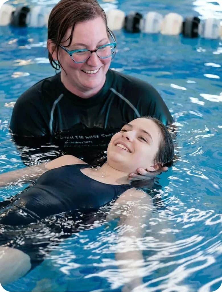 A smiling woman in glasses supports a young girl floating on her back in a swimming pool.