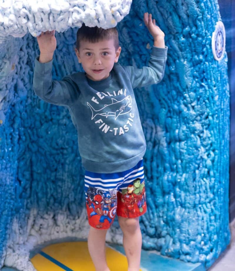 A young boy in a "FEELING FIN-TASTIC" shark shirt and superhero swim trunks stands under a blue wave.