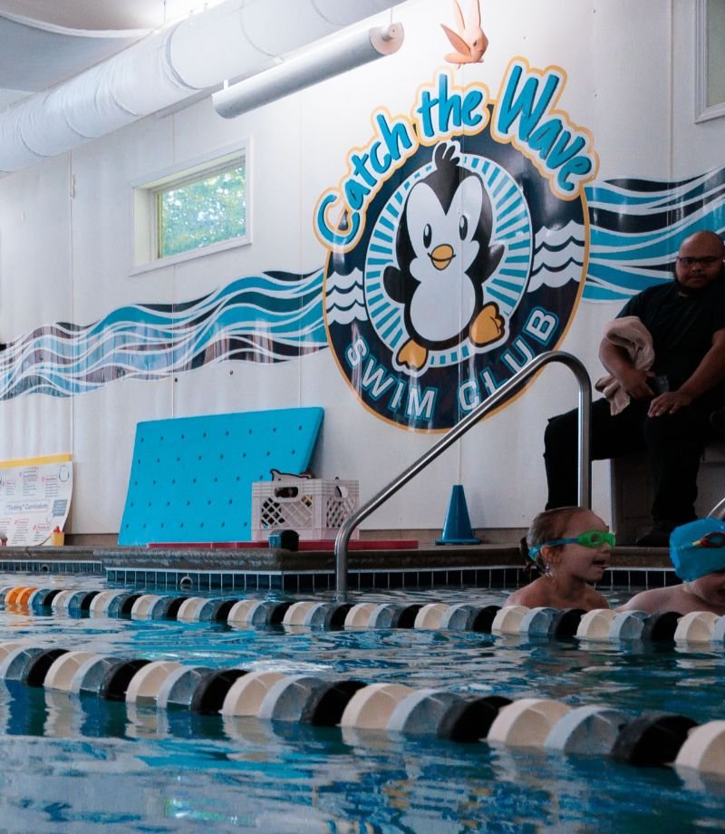 A girl in goggles swims in a pool with lane lines, under a "Catch the Wave Swim Club" penguin logo.