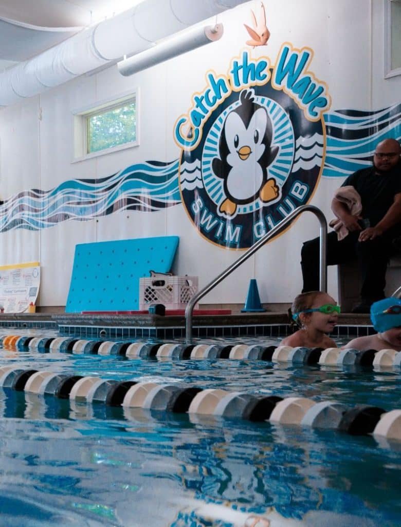 Two children in a swimming pool with lane dividers, a man watching, and a "Catch the Wave Swim Club" sign.