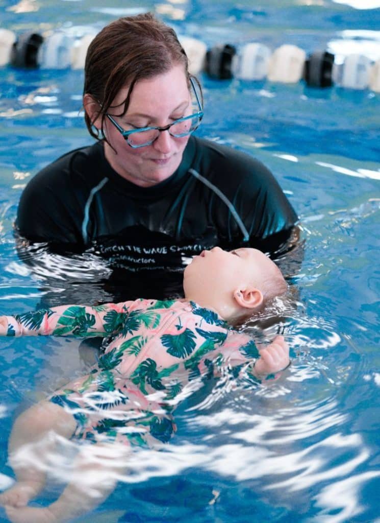 A woman in glasses and a black rashguard watches a baby in a pink and green swimsuit float on its back in a blue pool.