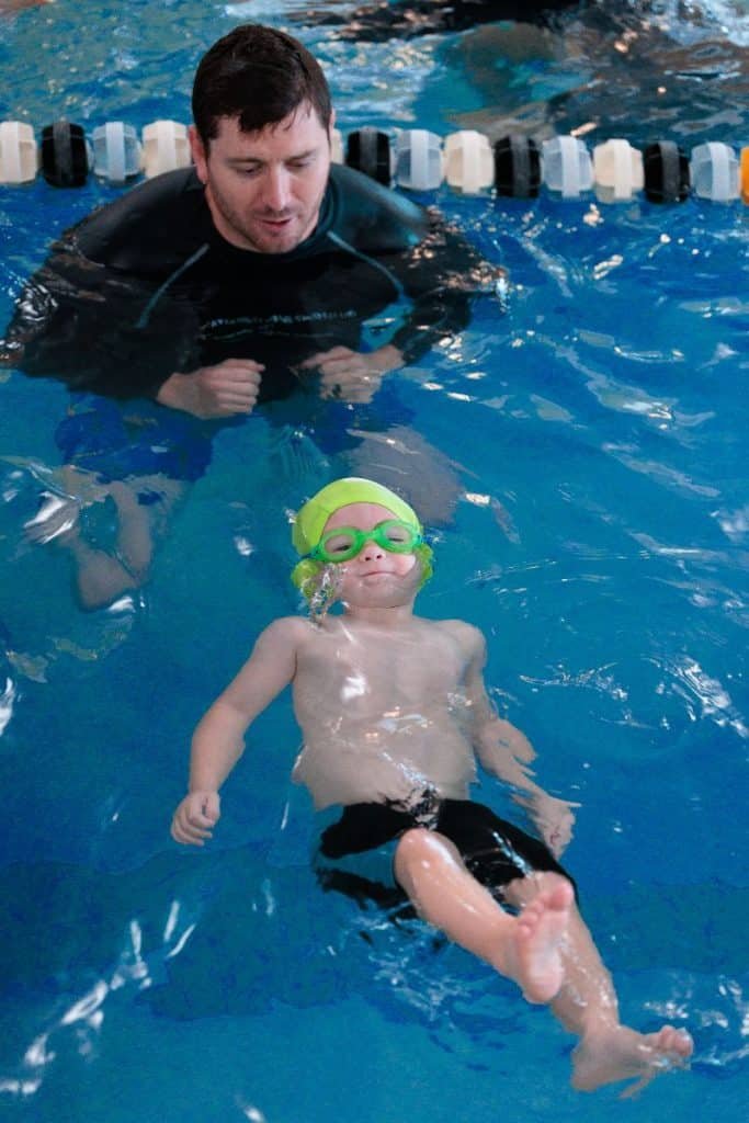 A man in a black shirt watches a child with a neon green swim cap and goggles floating on their back in a pool.