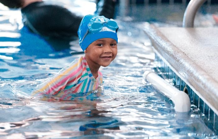 A smiling child in a blue swim cap and goggles looks at the camera while in a swimming pool.