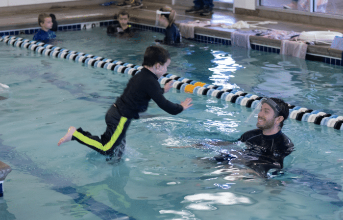 A child in a black and yellow wetsuit jumps into a pool towards a smiling man in a black wetsuit.