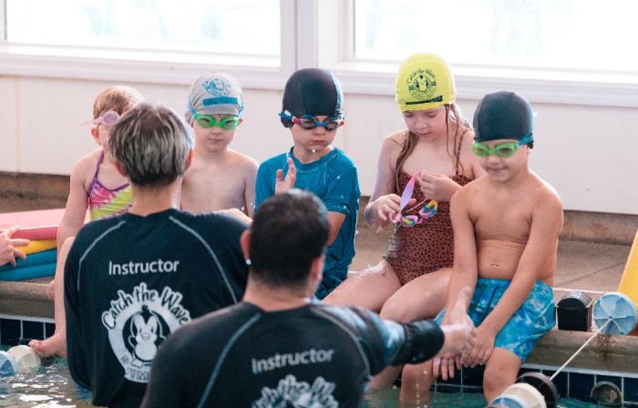 Two swim instructors in "Instructor" shirts face five children wearing swim caps and goggles at the pool's edge.