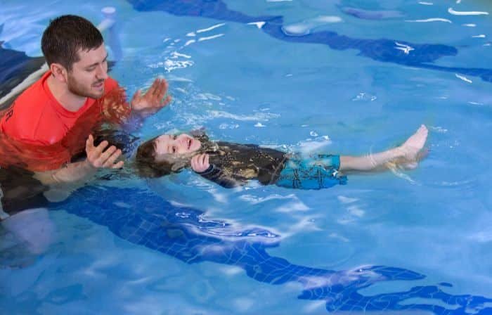 A man in a red shirt teaches a young child in a rashguard to float on their back in a blue pool.