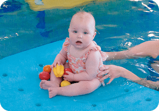 A baby in a swimsuit sits on a blue floating mat holding colorful toys in a swimming pool.