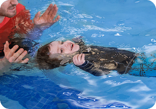 A smiling child in a black long-sleeved swim shirt floats on their back in a pool, supported by an adult.