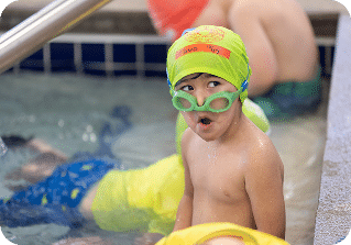 A young boy in a neon green swim cap and goggles stands in a swimming pool, looking right.