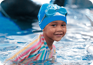 A smiling child in a blue swim cap, goggles, and a striped rash guard stands in a swimming pool.