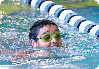 A young boy with dark hair and yellow goggles swims in a pool with a black and white lane rope.