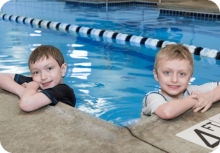 Two young boys with wet hair smile while leaning on the edge of a blue indoor swimming pool.