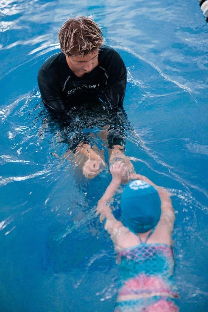 A blonde man in a black "CATCH THE WAVE SWIM" shirt teaches a child in a blue swim cap to float.