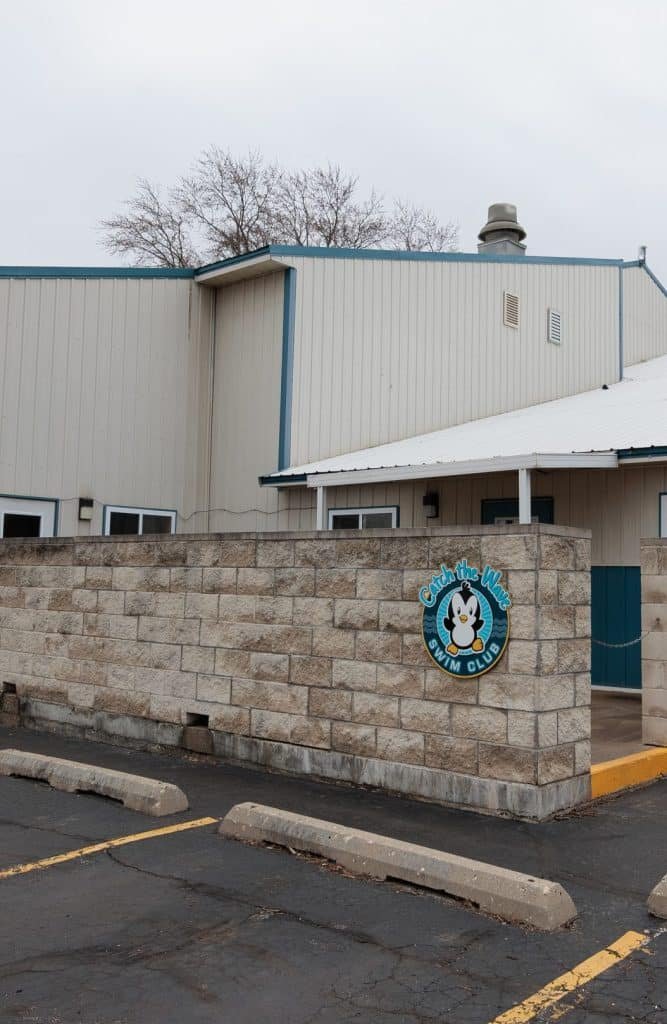 A "Catch the Wave Swim Club" sign with a penguin is on a stone wall next to a building and parking lot.