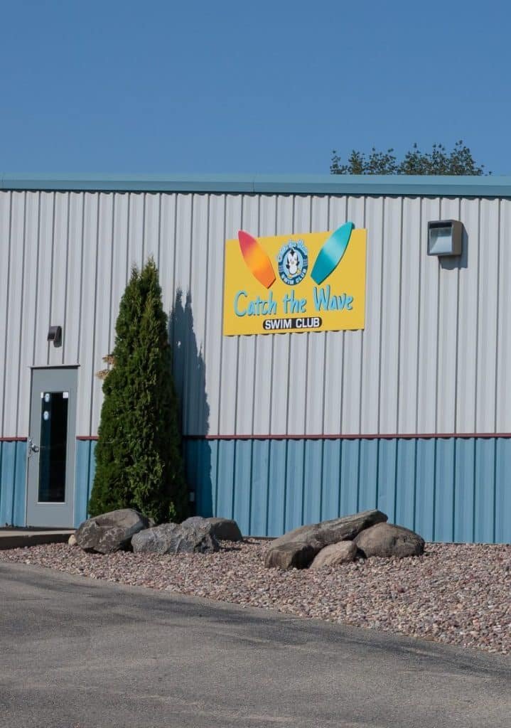 A white and blue corrugated building with a "Catch the Wave Swim Club" sign, a door, and landscaping.