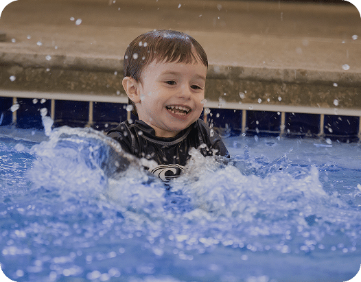 A young boy with short brown hair smiles widely, splashing in a blue swimming pool with water spraying around him.