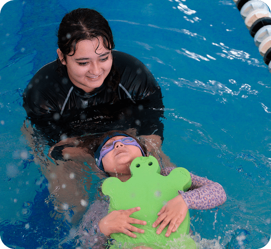 A smiling woman in a black rashguard supports a child wearing goggles and holding a green frog float in a pool.