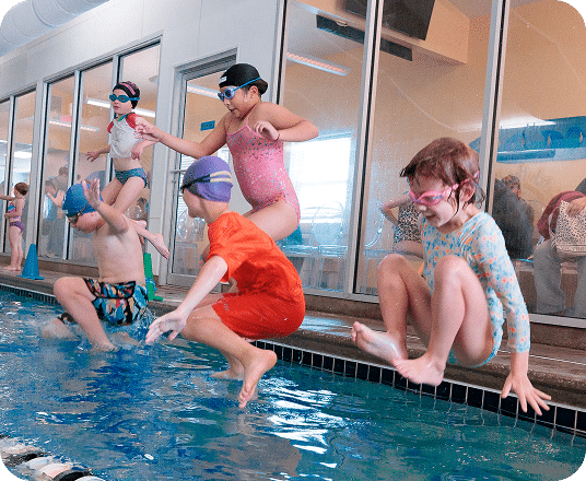 Five children in swimsuits and goggles jump into a blue indoor swimming pool.