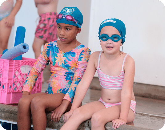 Two young girls in swimsuits, swim caps, and goggles sit on a pool edge next to a pink crate with blue noodles.