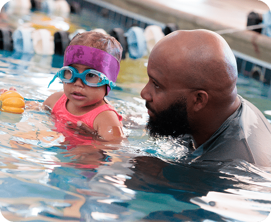A bald man with a beard looks at a child wearing a purple swim cap and blue goggles in a swimming pool.