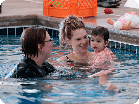 A woman in glasses and a smiling woman holding a baby are in a swimming pool.