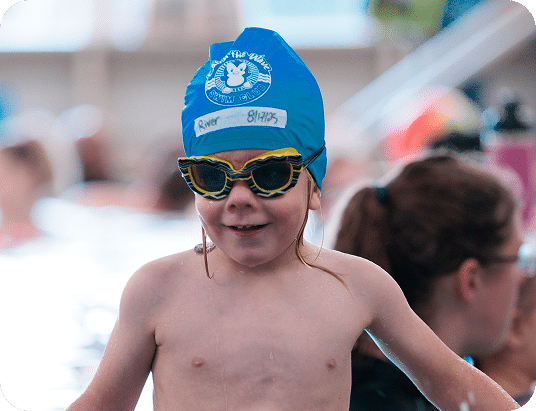 A smiling child in a blue swim cap with "River 8/17/23" and "Keep The Wave Swim Club" written on it, wearing striped goggles.