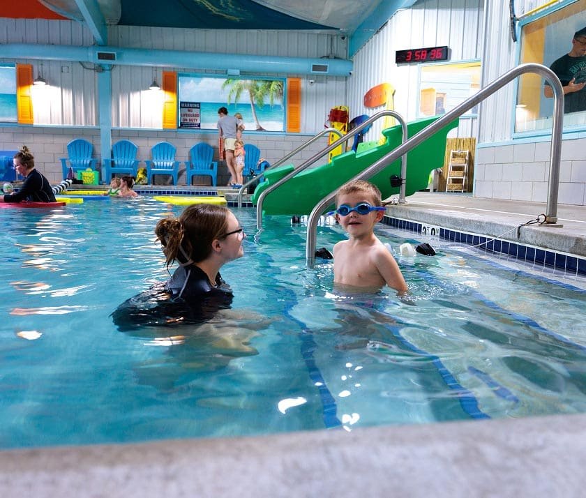 A young boy wearing goggles smiles at the camera while standing in a pool with an adult nearby.