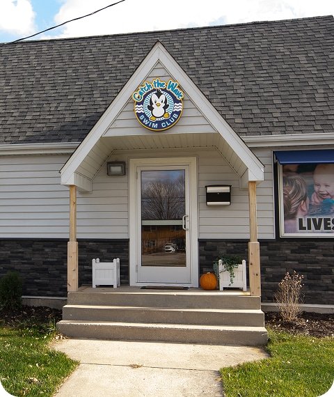 A building with a "Catch the Wave Swim Club" sign, a glass door, and concrete steps with a pumpkin.