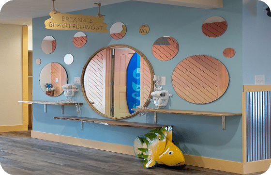 A light blue wall with a "BRIANA'S BEACH BLOWOUT" sign, circular mirrors, shelves, and a yellow fish statue.