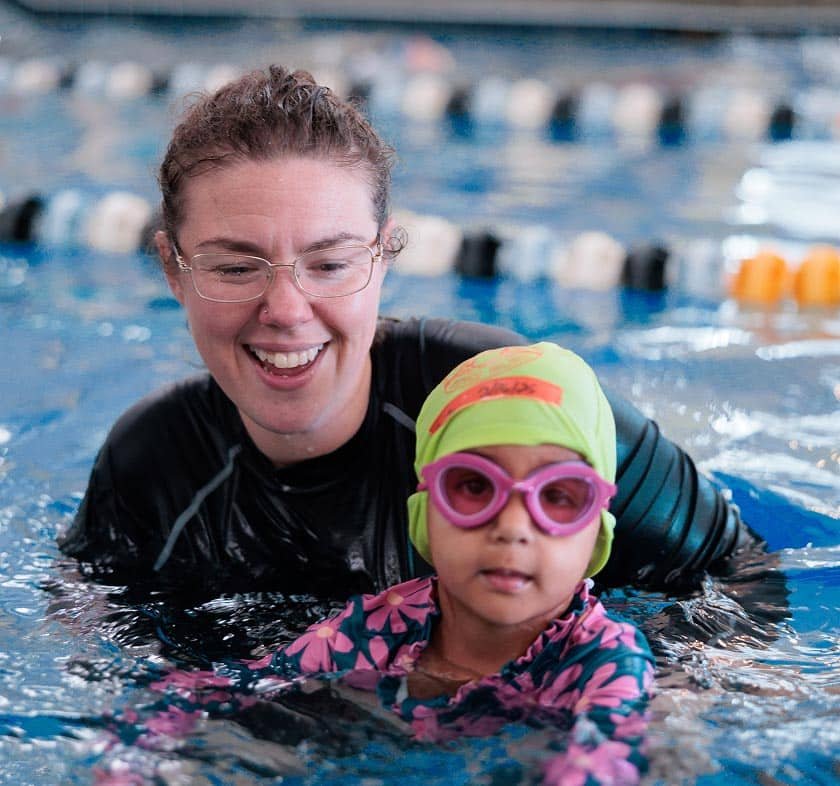A smiling woman in glasses and a black shirt supports a child in a green swim cap and pink goggles in a pool.