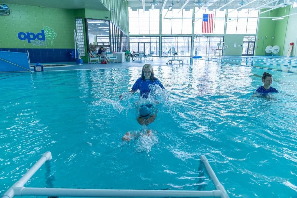 A smiling woman in a blue shirt splashes water while holding a child in a blue swim cap in a pool.