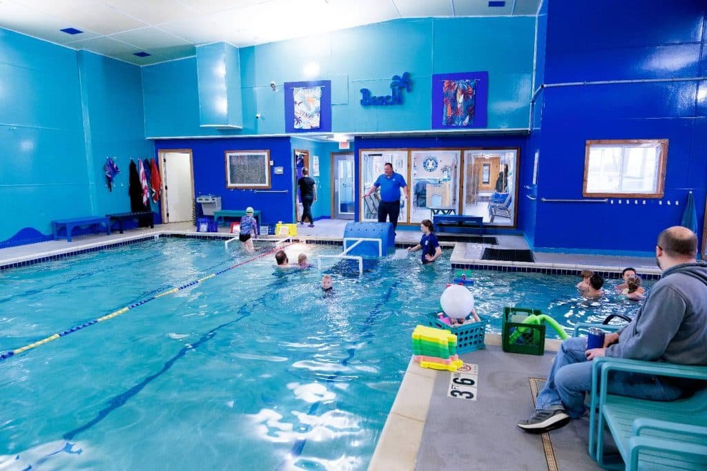 An indoor swimming pool with light blue walls, people swimming, and a man sitting poolside.