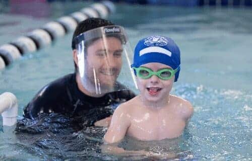 A smiling man wearing a "Flowfold" face shield teaches a young boy in a blue swim cap and green goggles to swim.