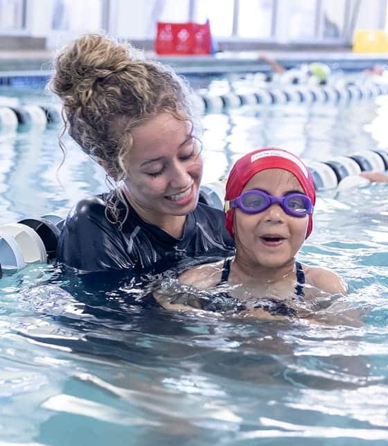A smiling woman with curly hair in a bun helps a young girl wearing a red swim cap and purple goggles in a pool.